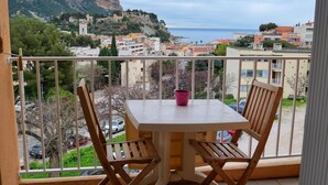 A balcony with a table and chairs, overlooking a coastal town with buildings and a mountain. - Apartment/ flat - Cassis (Cassis)