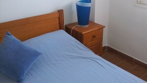 Une chambre à coucher avec un lit, une table de chevet en bois, un oreiller bleu et une poubelle bleue.