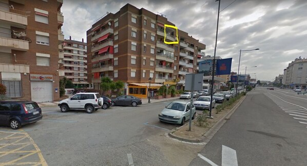 A multi-story residential building with balconies, a visible storefront, and parked cars along the street.