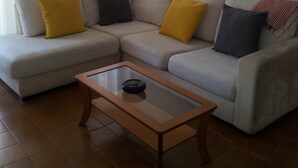 Un salon avec un canapé d’angle, une table basse en verre et une fenêtre avec des rideaux.