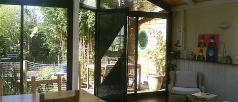 A room with a wooden table and chairs, a sofa, and a view of an outdoor area with a wooden deck and greenery.
