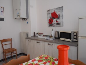 A kitchen with a microwave, a kettle, and a cabinet. There is a chair and a table with a floral tablecloth.
