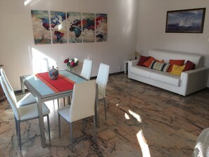A modern living room with a dining area, featuring a glass table, white chairs, a white sofa with colorful cushions, and a large abstract painting on the wall.