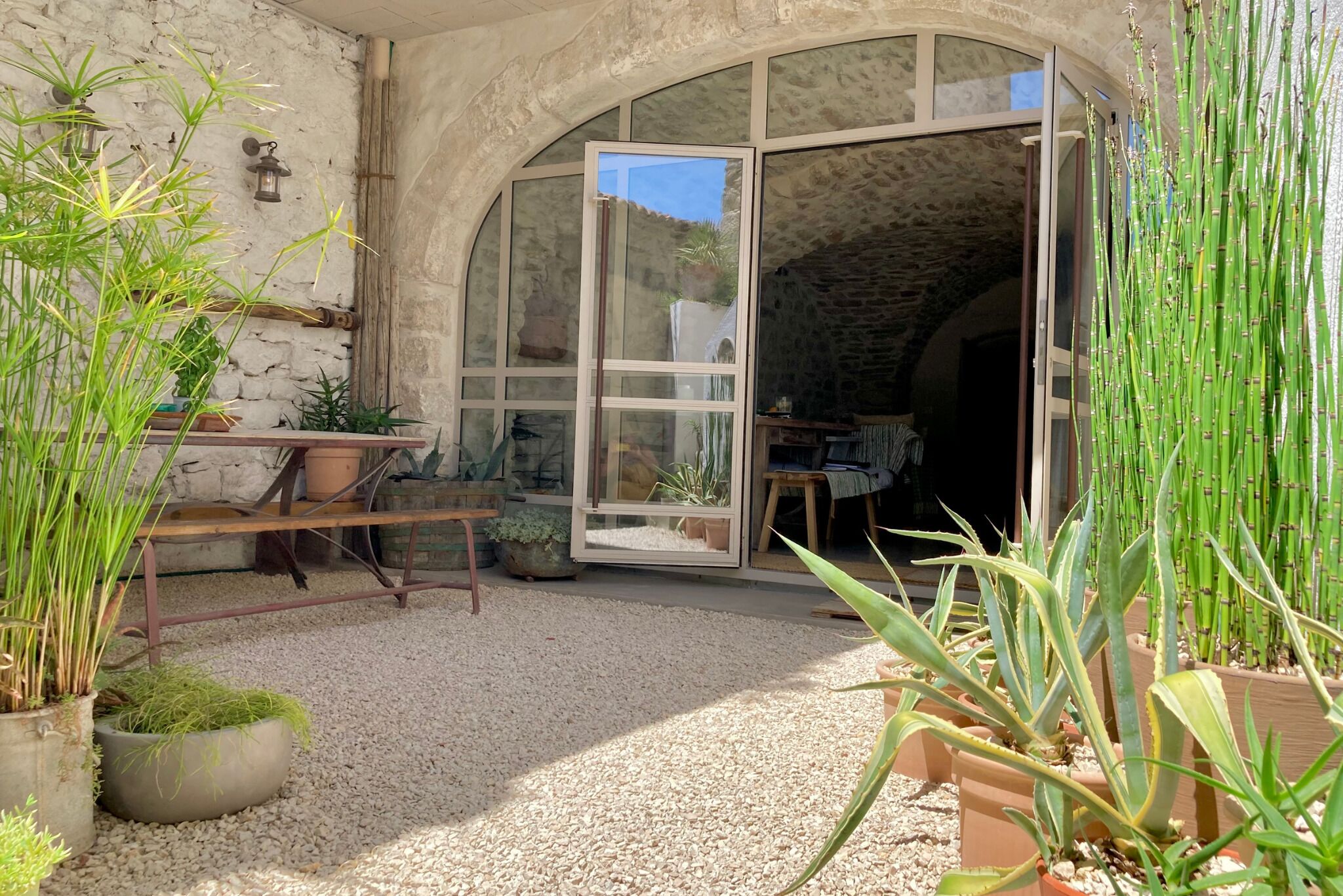 A rustic stone building with a large arched doorway leading to a room. There are potted plants and a wooden bench outside.