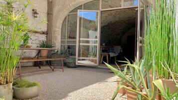 A rustic stone building with a large arched doorway leading to a room. There are potted plants and a wooden bench outside.