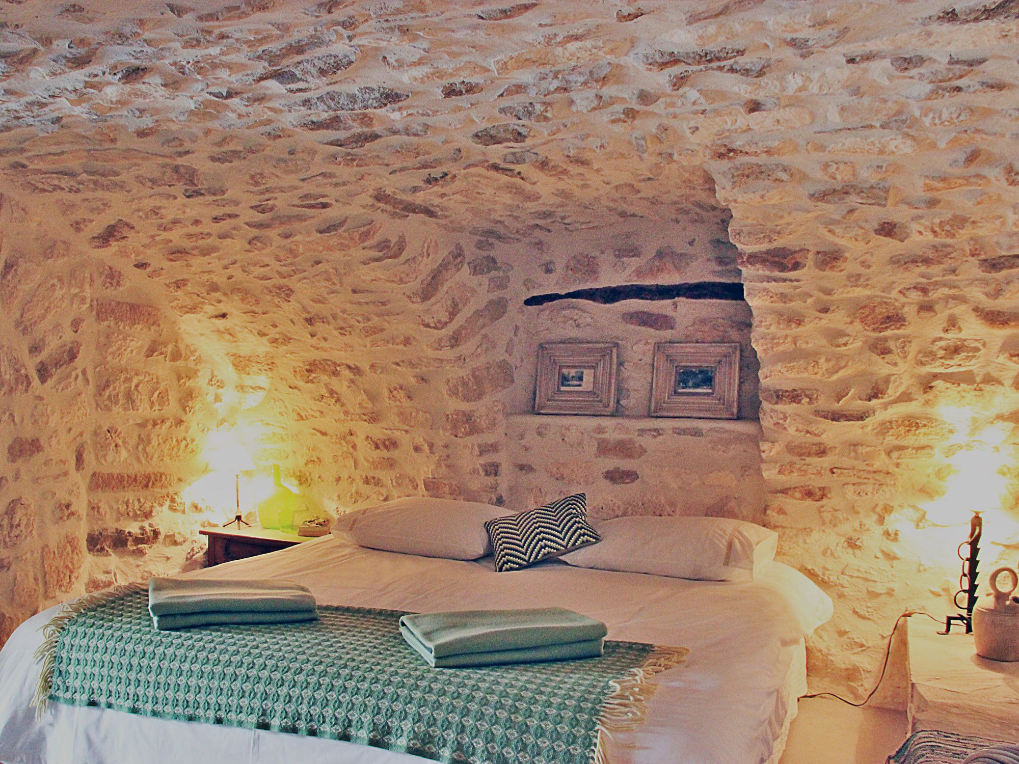 A bedroom with a stone wall, a bed with pillows, a green blanket, and a small table with a lamp.