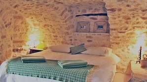 A bedroom with a stone wall, a bed with pillows, a green blanket, and a small table with a lamp.