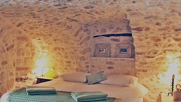 A bedroom with a stone wall, a bed with pillows, a green blanket, and a small table with a lamp.