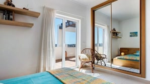 Une chambre avec une grande porte-fenêtre coulissante donnant sur un balcon, un lit recouvert d’une courtepointe bleue, une chaise et un grand miroir qui reflète la pièce.