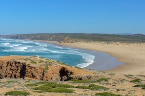 A coastal landscape with a sandy beach, rocky outcrops, and sparse vegetation.