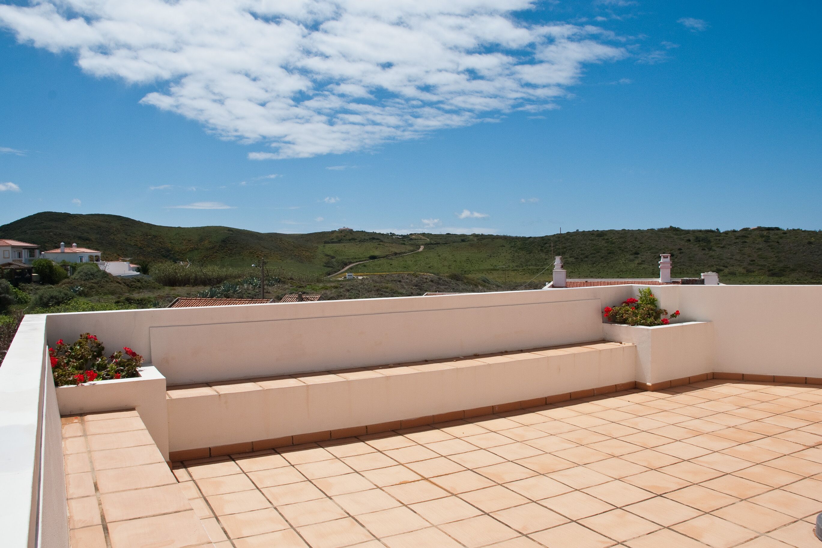 A rooftop terrace with a tiled floor, white walls, and flower planters.