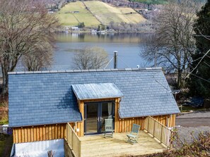 Une cabane en bois avec un toit sombre, une terrasse avec deux chaises et une vue sur un lac et des collines en arrière-plan.