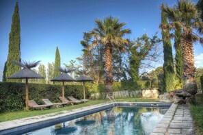 A swimming pool with lounge chairs and umbrellas, surrounded by tall trees and hedges.