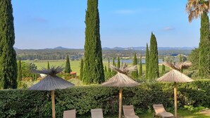 A landscaped outdoor area with lounge chairs and thatched umbrellas, surrounded by tall trees and a view of distant hills.