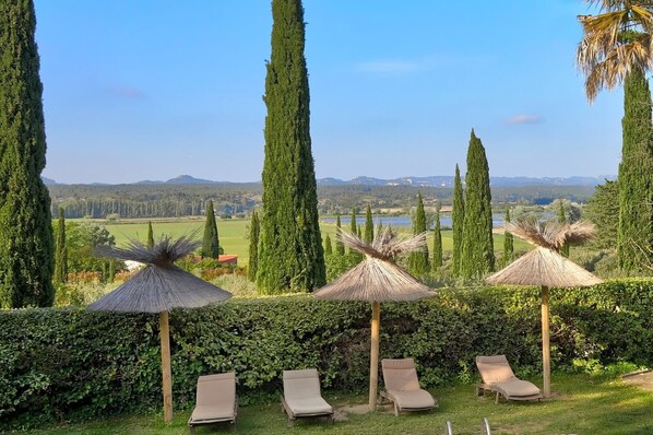 Un espace extérieur aménagé avec des chaises longues et des parasols en chaume, entouré de grands arbres et offrant une vue sur des collines lointaines.