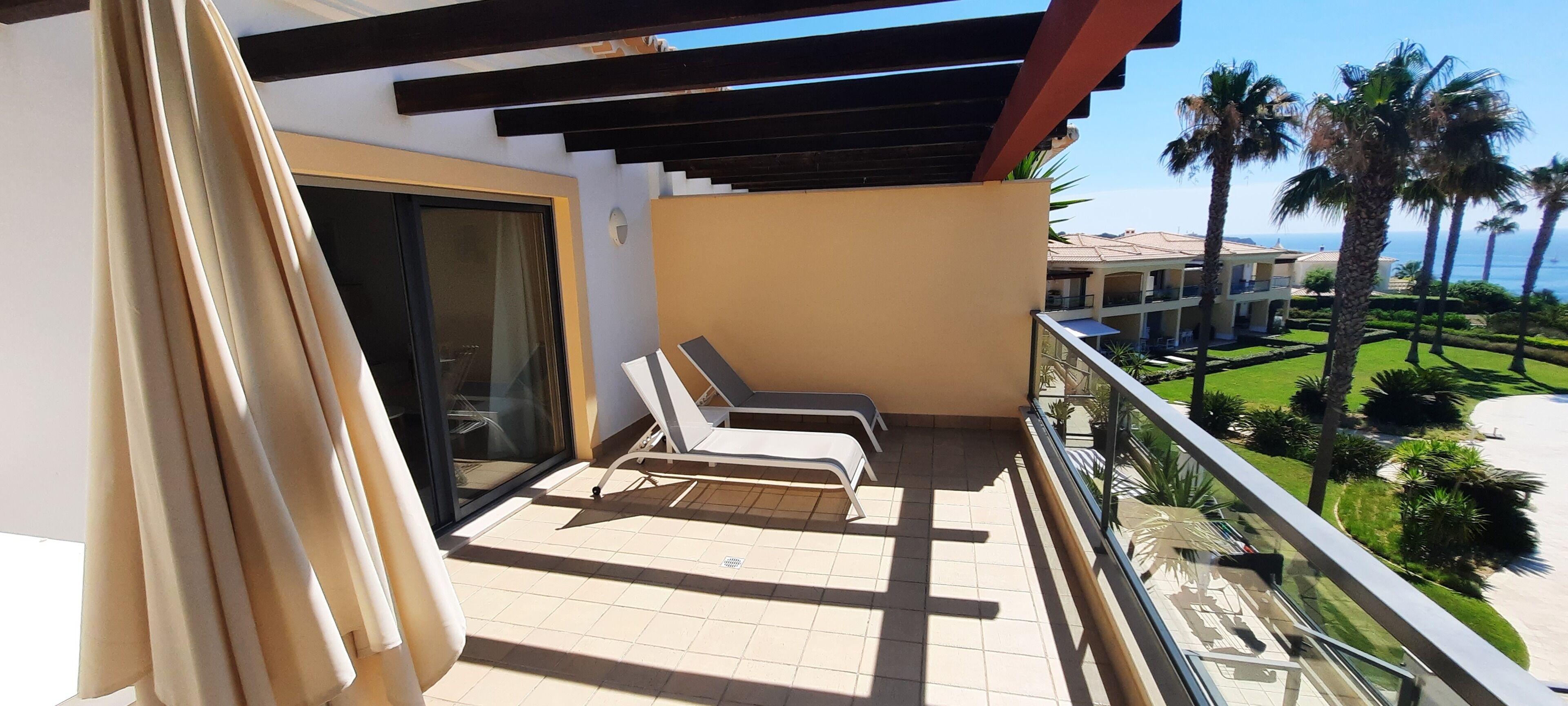 A balcony with two sun loungers, a glass door, and a view of a landscaped area with palm trees and a building in the background.