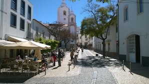 Una calle adoquinada bordeada de cafés con mesas al aire libre, una torre de iglesia al fondo y un camino peatonal.