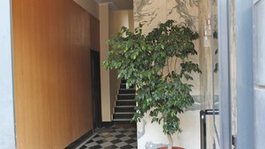 Een hal met een geruit vloerkleed, een potplant, een marmeren wand en een trap op de achtergrond.