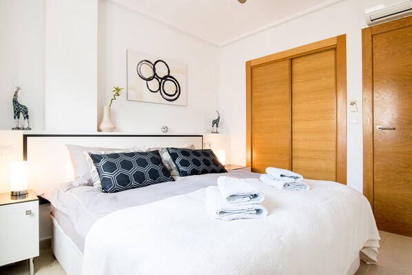 A neatly made bed with a white comforter and two patterned pillows, a bedside table with a lamp, a wall-mounted artwork, and a wooden wardrobe.
