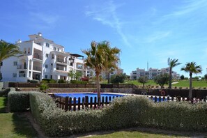 Een wit hotel met meerdere verdiepingen, balkons, een zwembad en palmbomen.