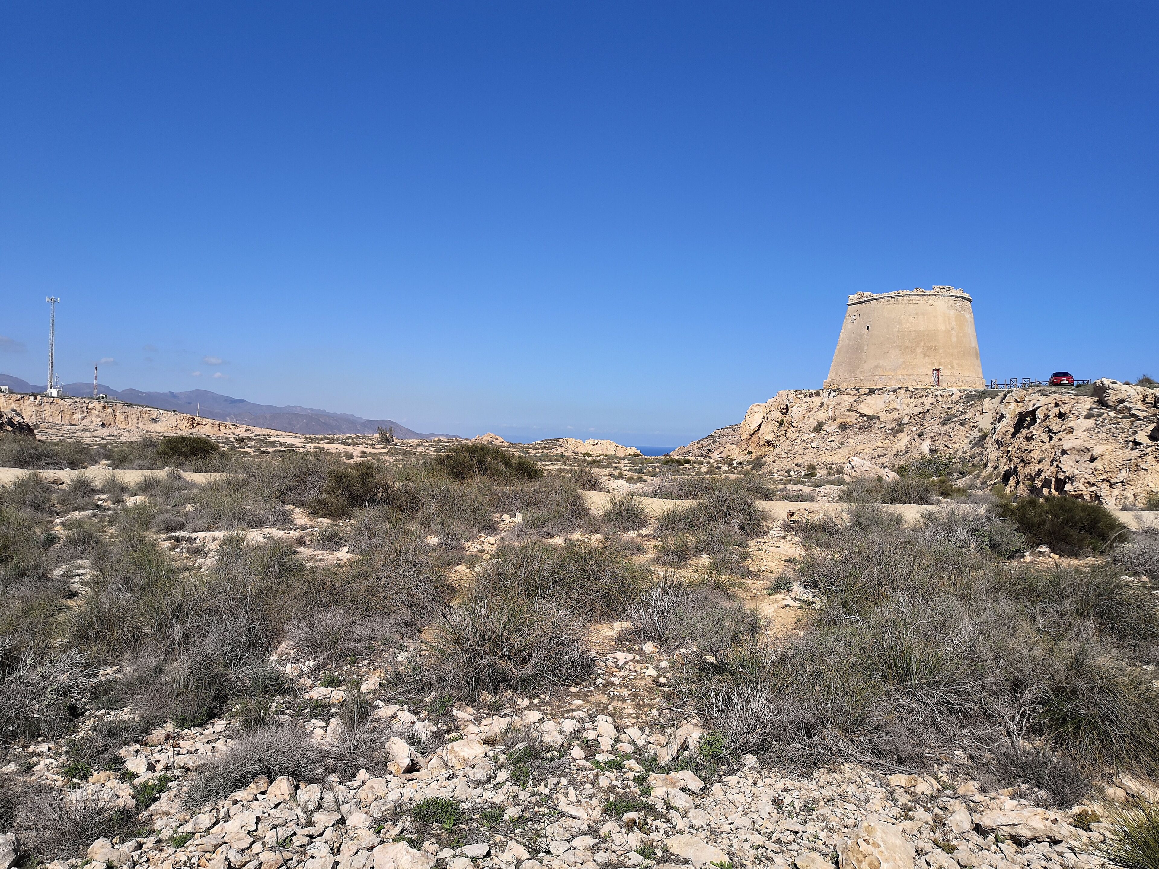 Paisaje desértico con una gran estructura de concreto en una colina, terreno rocoso, vegetación escasa y montañas al distante.