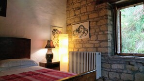 A bedroom with a bed, a nightstand with a lamp, a radiator, and two framed artworks on the stone wall.