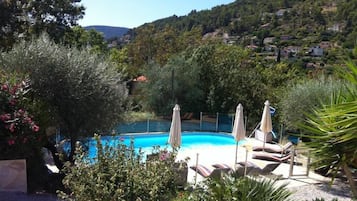 Una piscina con sillones de descanso y sombrillas, rodeada de vegetación y casas al fondo.