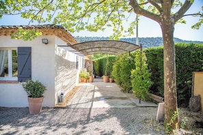 A covered outdoor area with a pergola, potted plants, and a gravel path leading to a building with a tiled roof.