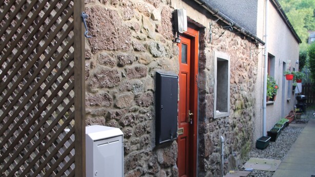 A stone building with a red door, a small window, and a utility box. - The Wee Hoose: Probably Scotland's Smallest Holiday Home  (Callander)