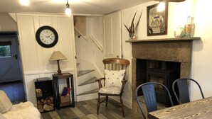 Une pièce chaleureuse avec une cheminée, une table à manger et une chaise. On y trouve un escalier avec un panneau d’avertissement, une horloge au mur et une lampe sur une table d’appoint.