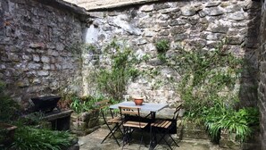 Une cour bordée de murs en pierre, avec une table et des chaises, entourée de verdure et agrémentée d’un banc en pierre.