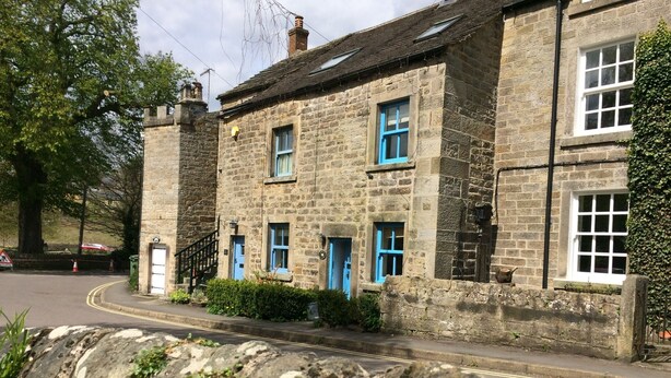 Un bâtiment en pierre avec des portes et des fenêtres bleues, un mur de pierre au premier plan et une route avec un panneau de signalisation.