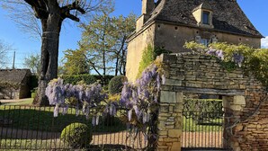 Un bâtiment en pierre doté d’un toit traditionnel, d’un grand arbre et d’une arche en pierre ornée de fleurs violettes.