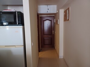 A narrow hallway with a white refrigerator on the left, a wooden door at the end, and a bulletin board on the right wall.