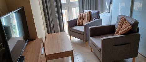 A modern living room with a wooden table, two grey sofas with striped cushions, a large window with curtains, and a laptop on the table.