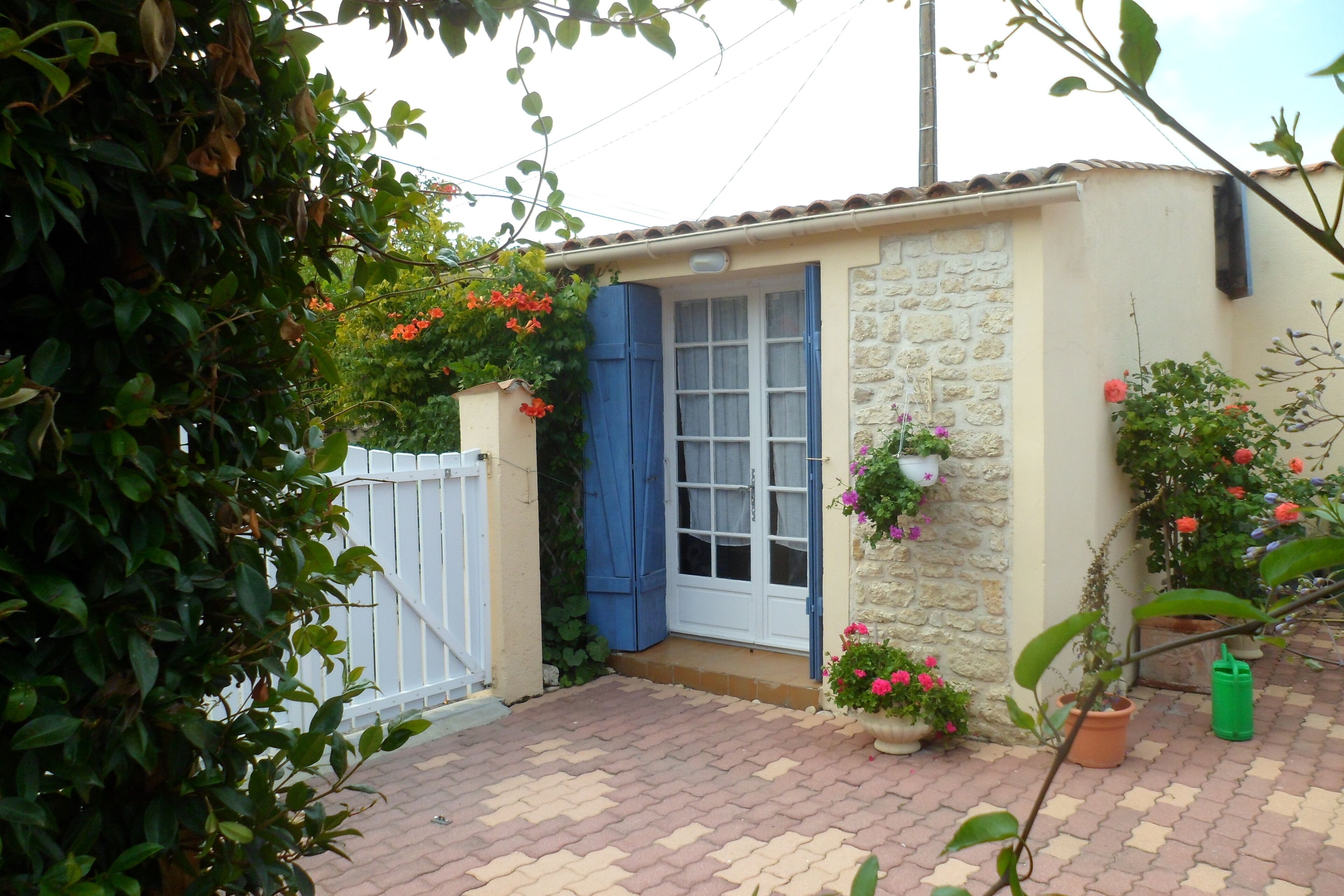 A small, stone building with a blue door and windows, surrounded by potted plants and a brick pathway.