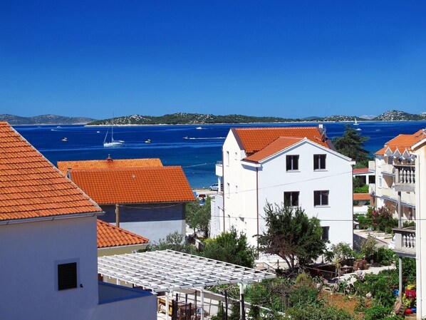 A coastal town with white buildings and orange roofs, a clear blue sea, and boats in the distance.