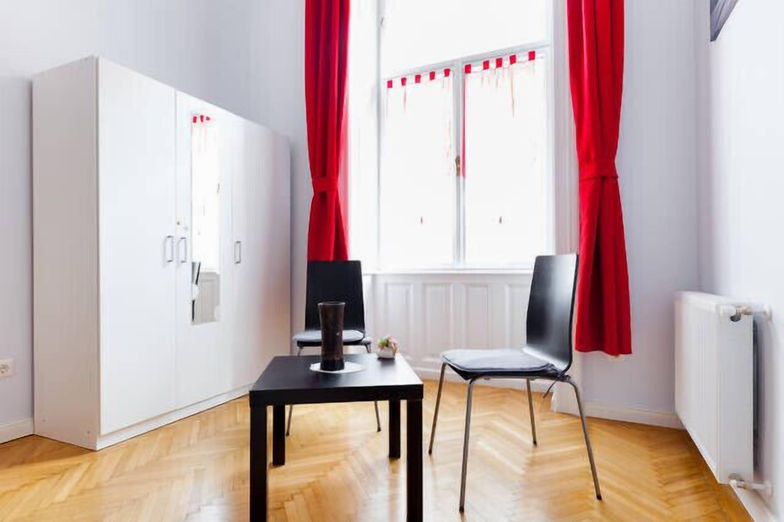 A room with a white wardrobe, a black table with a vase, a black chair, and red curtains.