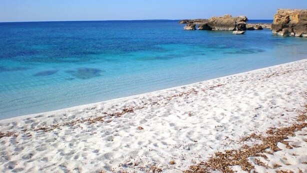 A beach with clear blue water and white sand, featuring rocky formations in the background.