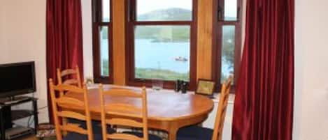 A dining area with a wooden table and chairs, a television, and a view of a lake through large windows.