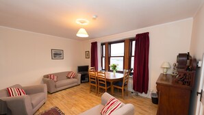 A living room with two sofas, a dining table with chairs, a TV, and a wooden cabinet. - Detached spacious Isle of Lewis cottage in tranquil position with loch view (Carloway)