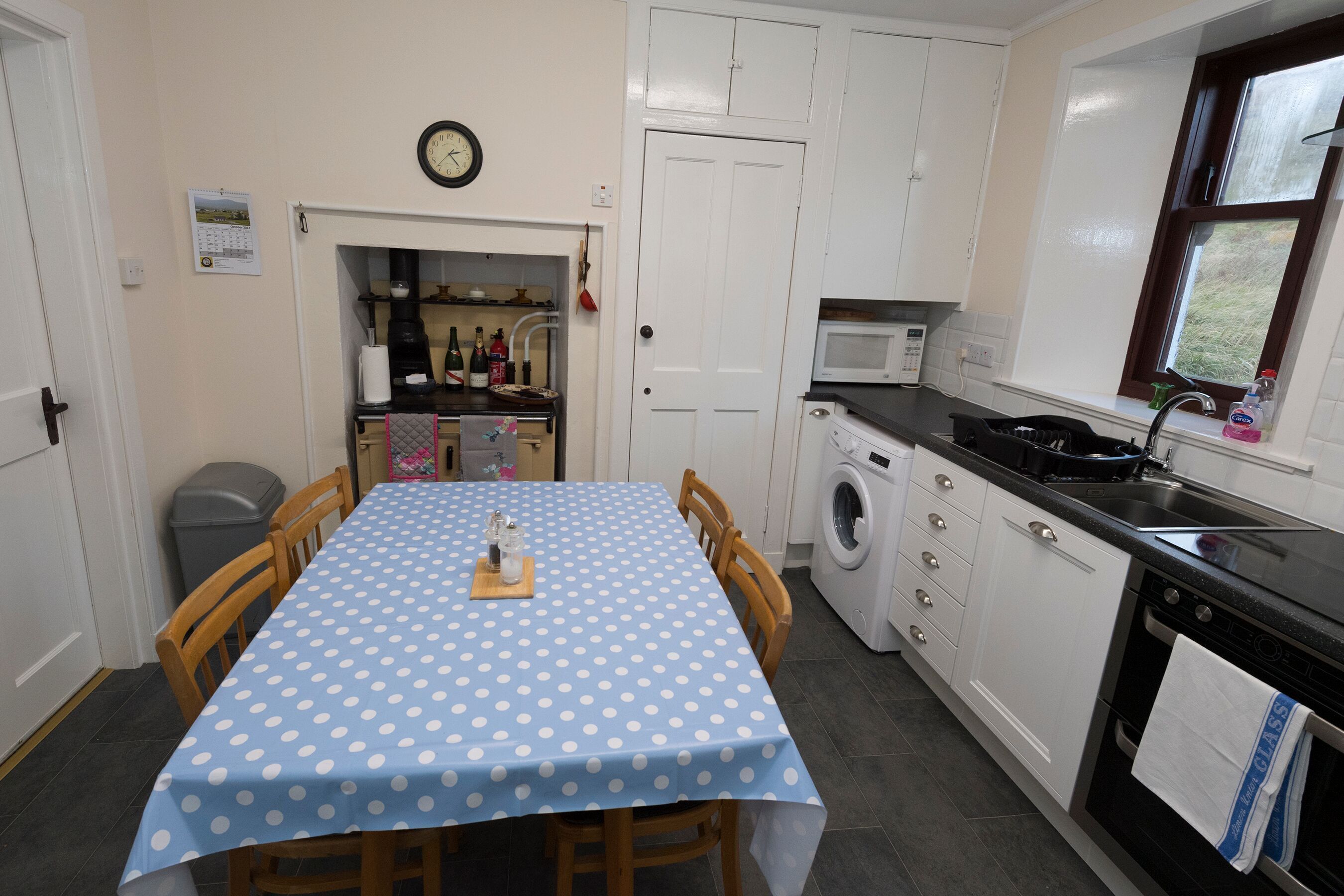 A kitchen with a blue polka dot tablecloth, white cabinets, a microwave, a washing machine, and a clock on the wall.