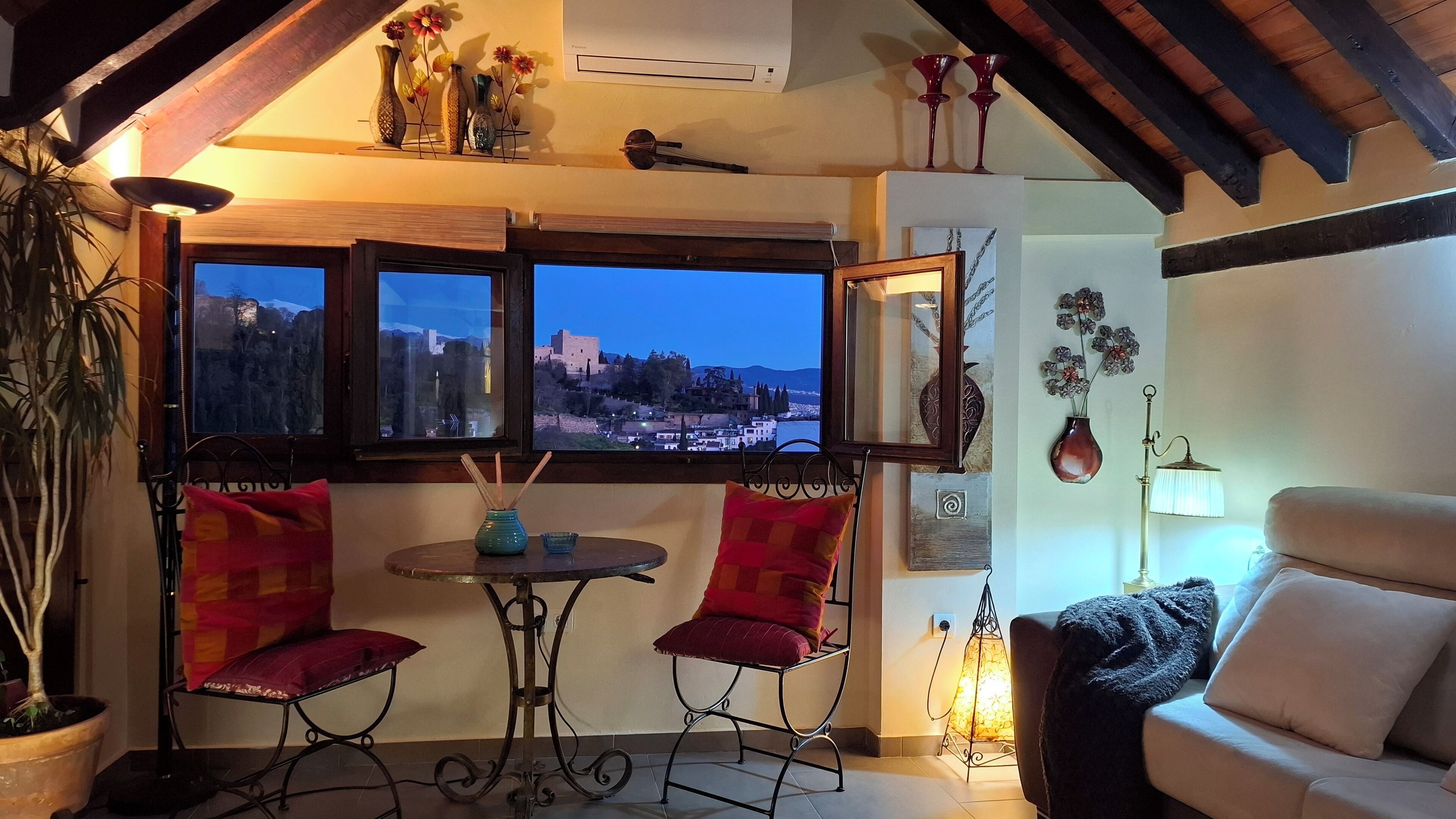 Une pièce chaleureuse donnant sur un paysage urbain au crépuscule, avec une table et deux chaises, un canapé et des objets décoratifs accrochés aux murs.