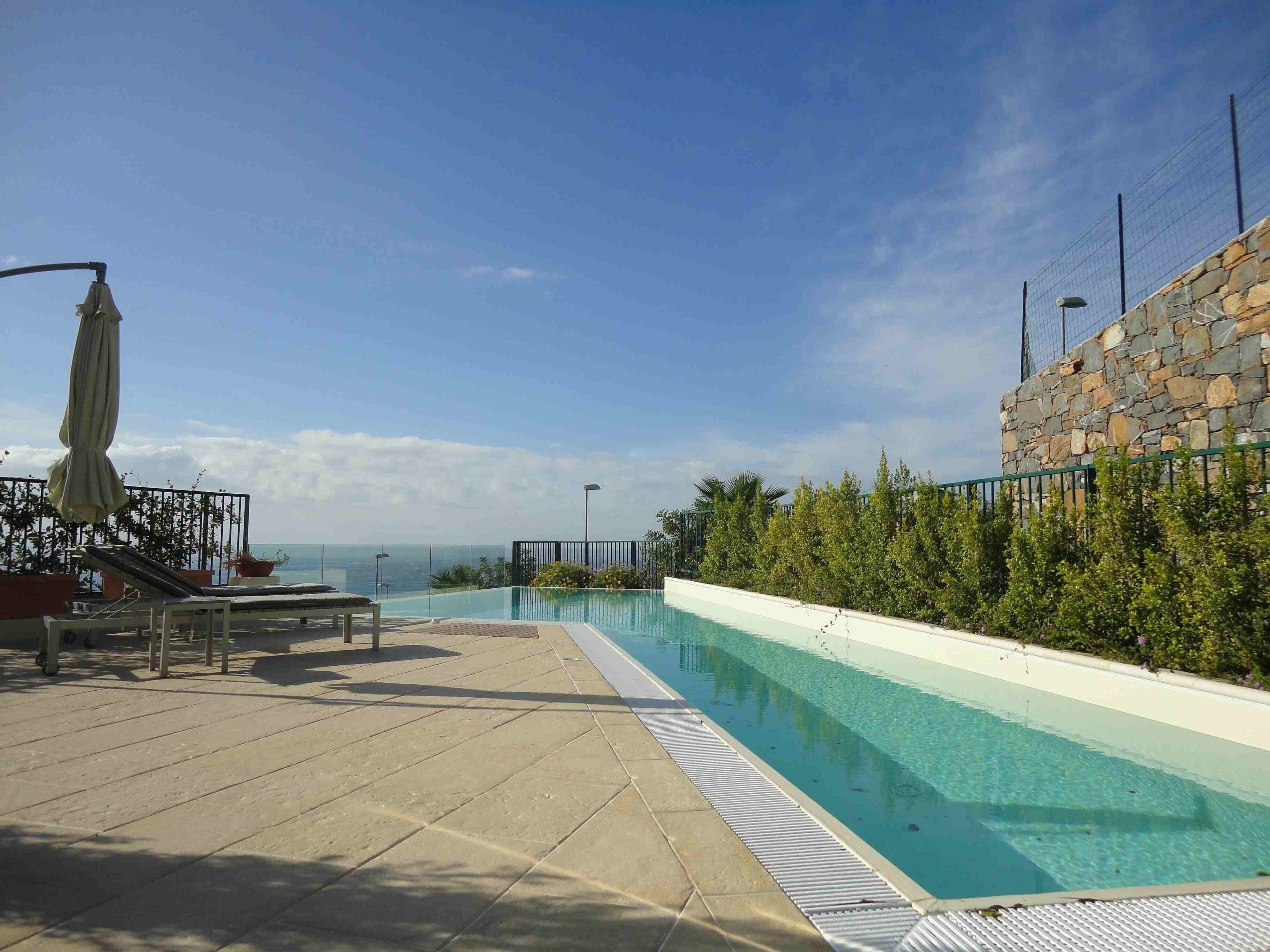 Blick auf einen Pool mit einer Steinmauer und viel Grün auf der einen Seite, und einer Liegebereich mit Liege und Sonnenschirm auf der anderen Seite.