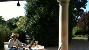 Une femme est assise à une table en plein air, recouverte d’une nappe blanche, et est entourée de verdure et d’une colonne en pierre.