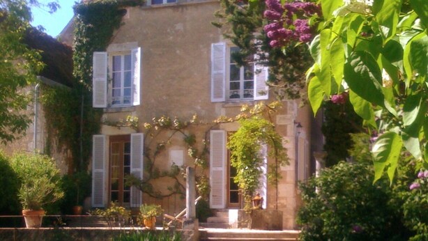 Un bâtiment en pierre avec des volets blancs, un jardin avec des marches menant à l’entrée et des plantes en fleurs.