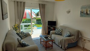 A living room with two patterned sofas, a coffee table, a TV, and a view of a pool and garden through a glass door. - Lovely Apartment, Ponta da Piedade Natural Reserve, Camilo Beach, Dona Ana Beach (Lagos)