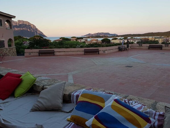 A terrace with a view of the sea and mountains, featuring a seating area with cushions and a blanket.