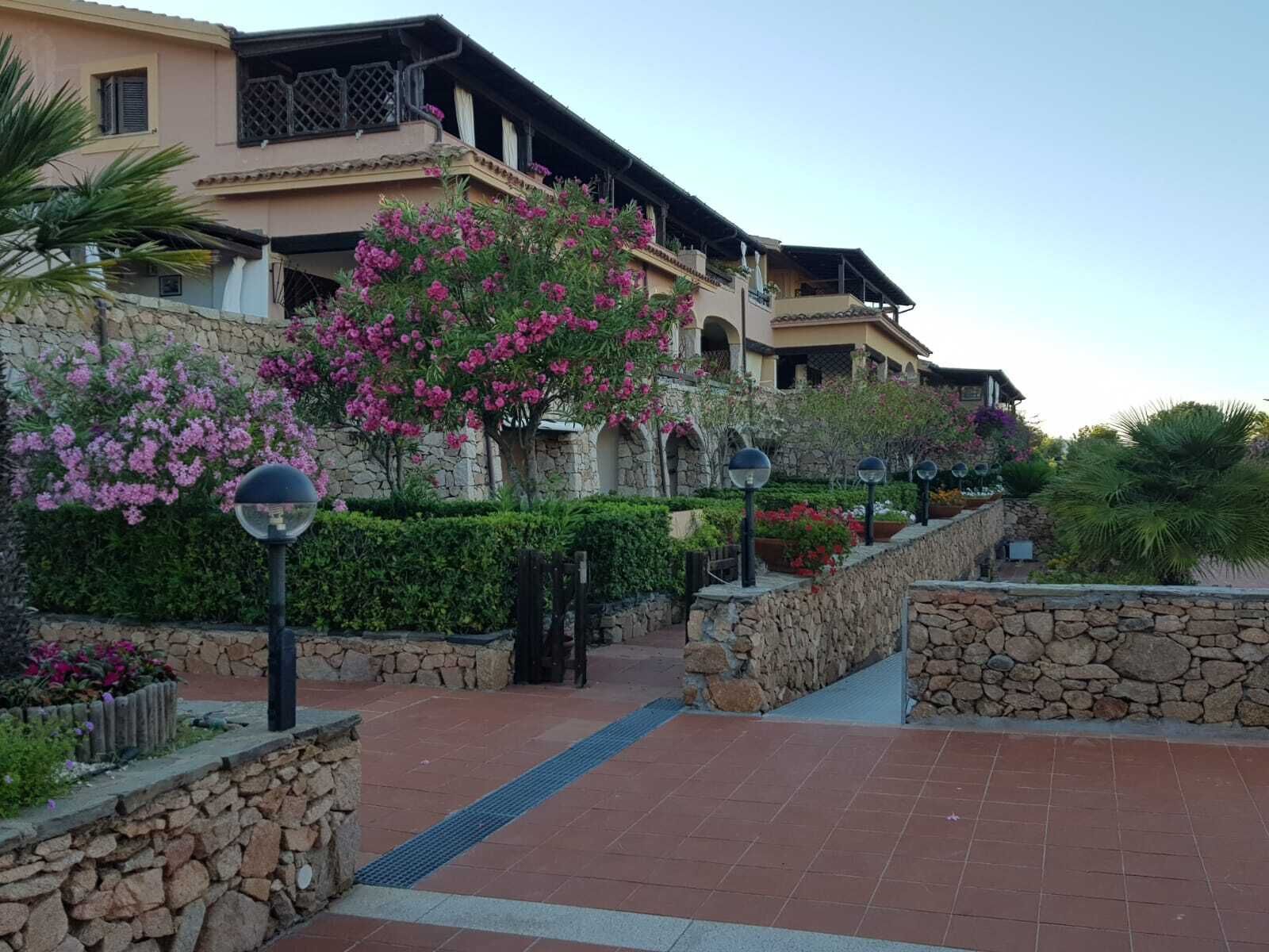Hotel amb façana de pedra, un camí pavimentat i una varietat de plantes de flors.