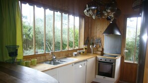 A kitchen with wooden cabinets, a sink, a stove, and a window overlooking greenery. - Cap Ferret charming wooden villa (Lège-Cap-Ferret)